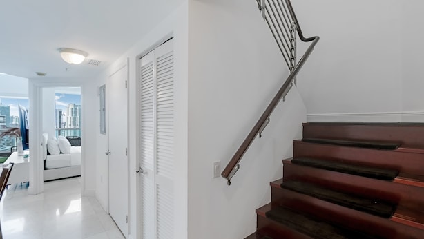 Un escalier avec une rampe, des murs blancs et une vue sur la ville par une porte ouverte.