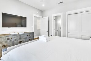 A modern bedroom with a large bed, a wooden dresser, a flat-screen TV, and white doors.