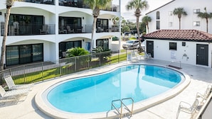 A swimming pool with a sun deck, surrounded by a white building with balconies and palm trees.