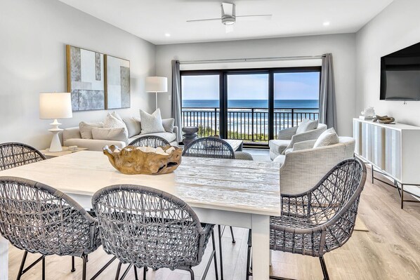 A modern living room with a large dining table, wicker chairs, a white sofa, and a view of the ocean.