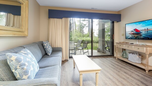 A living room with a sofa, a coffee table, a television, and a sliding glass door leading to an outdoor area with a deck and chairs. - Beautiful 2 Bedroom Lakeside Condo at The Shores 5677 (Williamsburg)