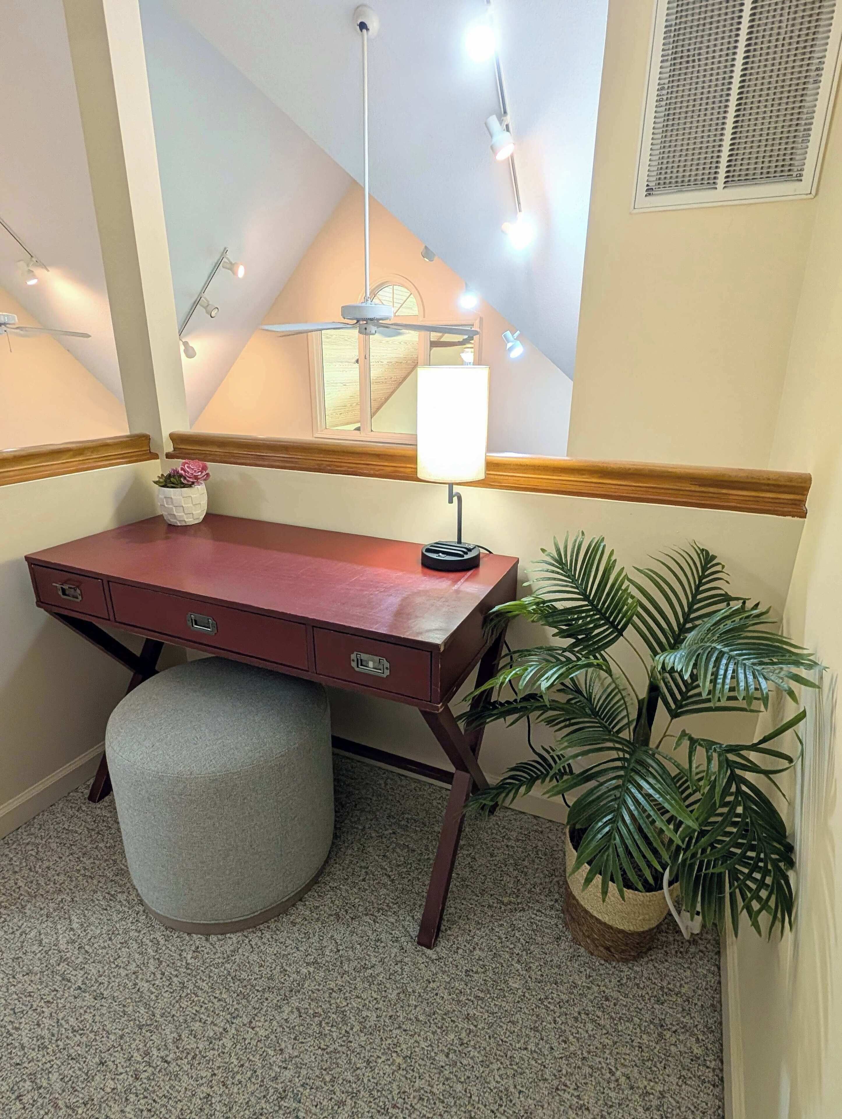 A desk with drawers, a lamp, a potted plant, and a ceiling fan in a room with a sloped ceiling and a window.