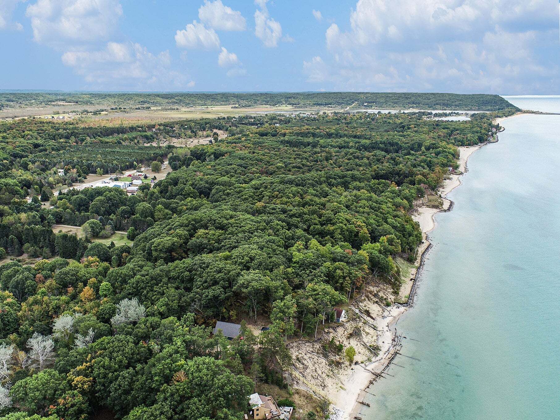 Luftaufnahme einer Küstenlandschaft mit dichtem Wald, einem Strand und verstreuten Gebäuden.