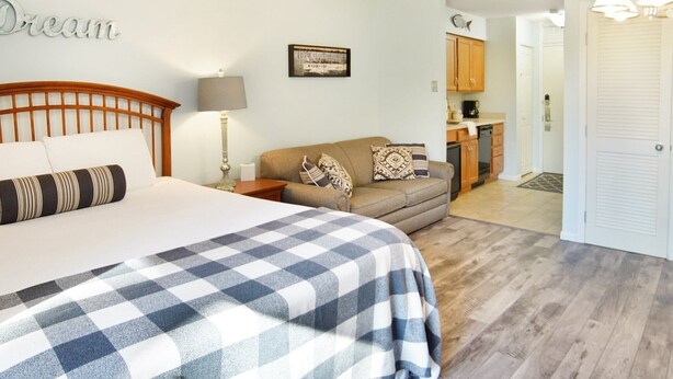 Een slaapkamer met een groot bed, een slaapbank, een nachtkastje, een lamp, een ingelijst kunstwerk aan de muur, en een keuken die zichtbaar is door een geopende deur.