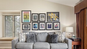 A living room with a grey sofa, framed pictures on the wall, a window with blinds, and a lamp on a side table.