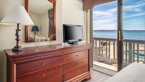A wooden dresser with a television, a lamp, and a mirror in a room with a balcony overlooking a beach.