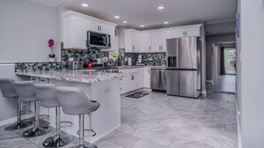 A modern kitchen with white cabinets, a stainless steel refrigerator, a microwave, and a marble countertop. There are three bar stools and a small island with a kettle. - The Riverwalk Retreat on the Fox - Waterfront Vacation Rental in Downtown Mchenry 3br/2ba Home With Private Pier (McHenry)