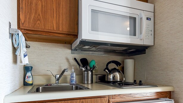 A compact kitchen with wooden cabinets, a white microwave, a sink with utensils, and a small refrigerator. - Lovely Beachside Studio 5679 (Williamsburg)