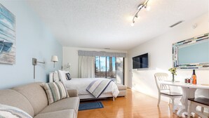 A modern bedroom with a bed, a sofa, a dining table with chairs, and a large window with curtains. - Lovely Beachside Studio 5679 (Williamsburg)