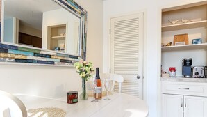 A white dining table with a vase of flowers, a bottle, and two glasses. A mirror hangs above the table, and a white door is visible in the background. - Lovely Beachside Studio 5679 (Williamsburg)