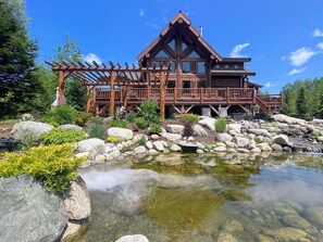 A lodge with a wooden deck, a pond with clear water, and surrounding greenery. - 1800wm WM Dream Home in National Forest (Bretton Woods)