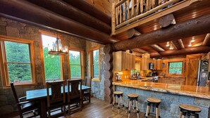 Interior rústico de una cabaña con pared de piedra, mesa de comedor de madera, taburetes de bar y una zona de cocina con armarios de madera y un ventanal grande.