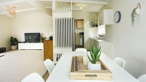 A modern living room with a TV, white furniture, a wooden divider, and a dining area with a tray. - Coastal Haven, 5 minute walk to beach. (Flinders)