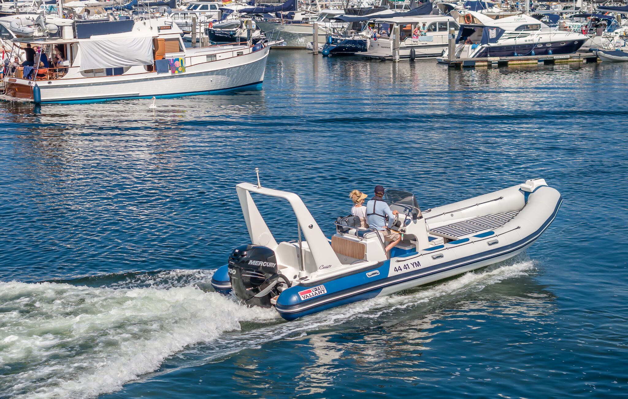 A motorboat with two passengers, moving through a marina with other boats docked.