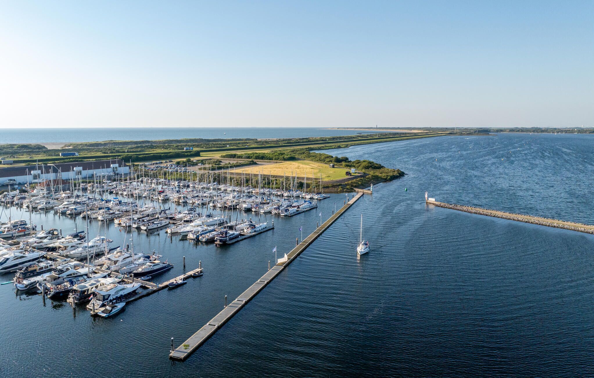 An aerial view of a marina with numerous boats docked, a long pier, and a distant shoreline with greenery.