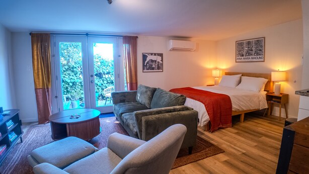 A hotel room with a bed, a sofa, a chair, a small table, and a view of greenery through the windows.