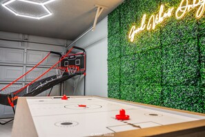 Een kamer met een tafelvoetbaltafel, een basketbalmand en een neonbord aan de muur.