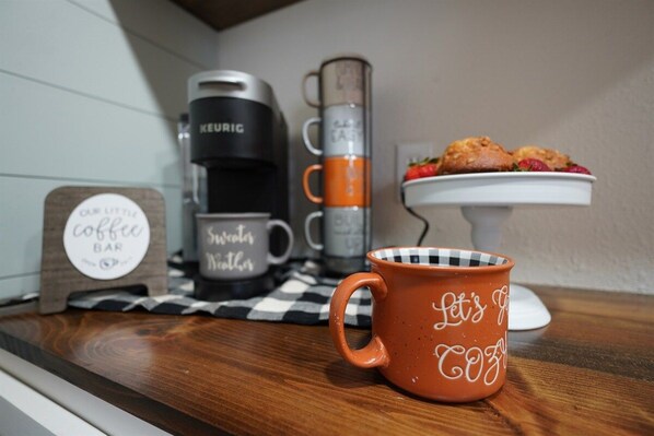 En köksbänk med en kaffebryggare, en hög med muggar, en skylt med texten ”Vårt lilla café” och en mugg med texten ”Låt oss gå”.