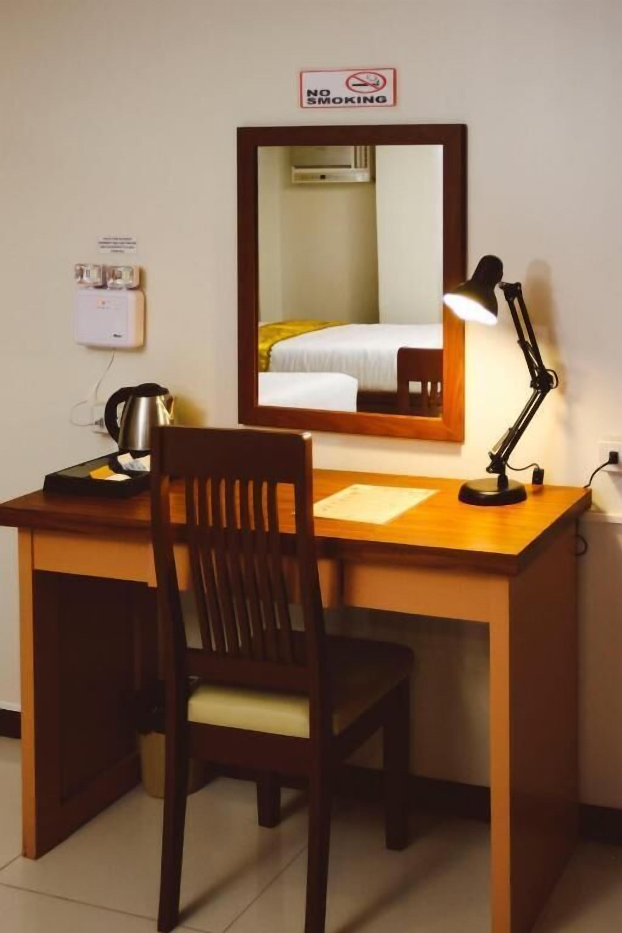 A hotel room with a wooden desk, a chair, a desk lamp, a kettle, and a bed with a white and yellow bedspread.