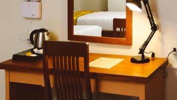 A hotel room with a wooden desk, a chair, a desk lamp, a kettle, and a bed with a white and yellow bedspread.