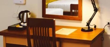 Habitación de hotel con un escritorio de madera, una silla, una lámpara de escritorio, un hervidor y una cama con cubrecama blanco y amarillo.