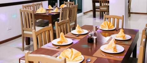A dining area with wooden tables and chairs, each set with folded napkins and cutlery.