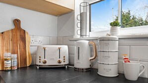 Una cocina con una tabla de cortar de madera, dos tostadoras blancas, una cafetera y una taza con una bolsita de té.