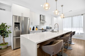 Cozinha moderna com geladeira de aço inoxidável, dispenser de água, fogão e pia. Há banquetas e uma janela grande com cortinas.