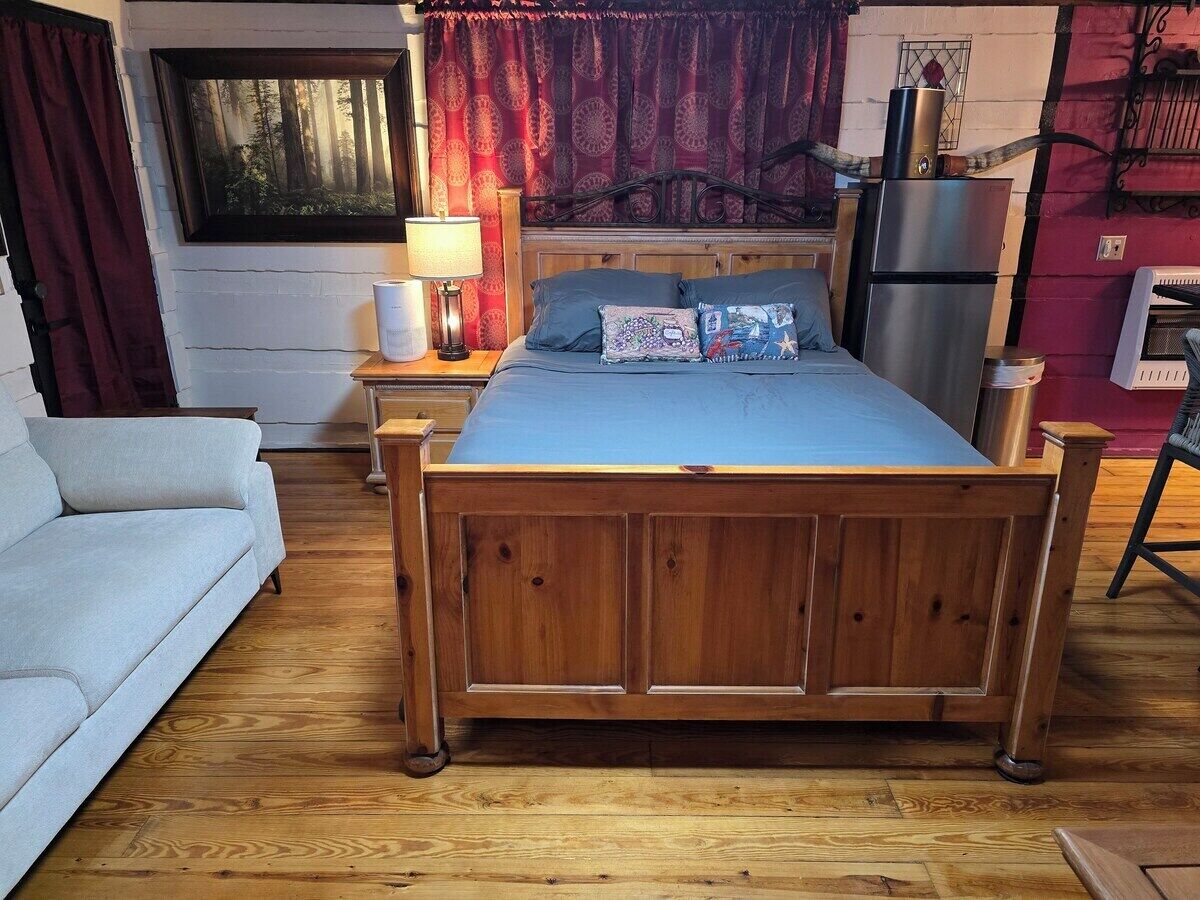 Un dormitorio con cama de madera, sofá blanco, mesita de noche con lámpara, refrigerador y ventana con cortinas.