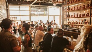 Um bar com clientes sentados em mesas, diversos garrafas nas prateleiras e uma parede de tijolos.