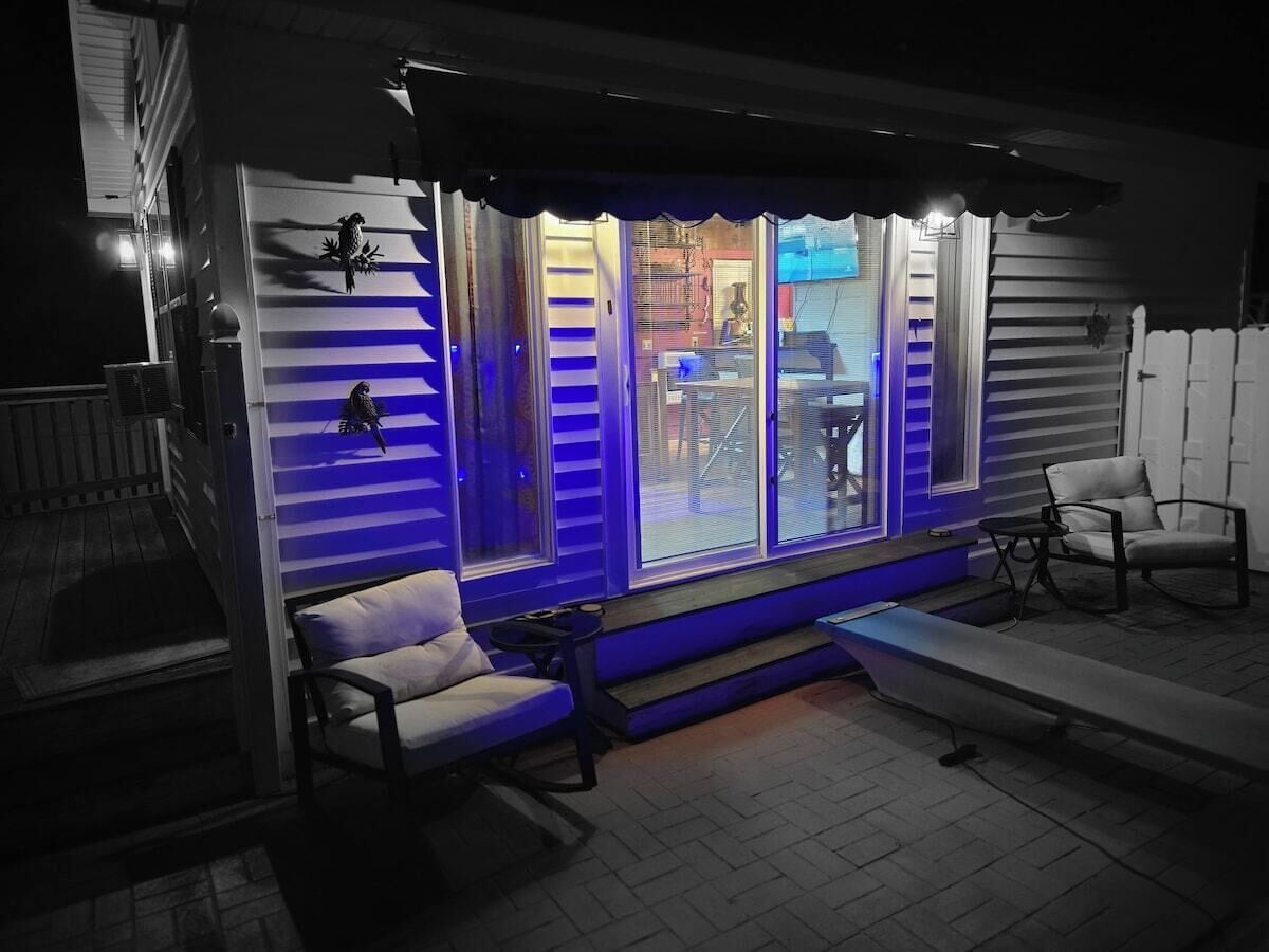 A covered outdoor seating area with a bench and chairs, a small table, and a window with blue lighting.