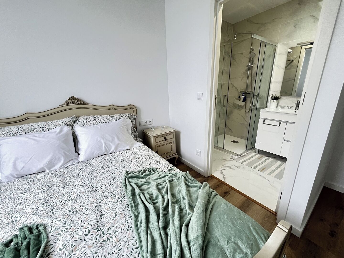 Ein Schlafzimmer mit einem Bett, einem Nachttisch, einer grünen Decke und einem sichtbaren Duschbereich durch eine offene Tür.