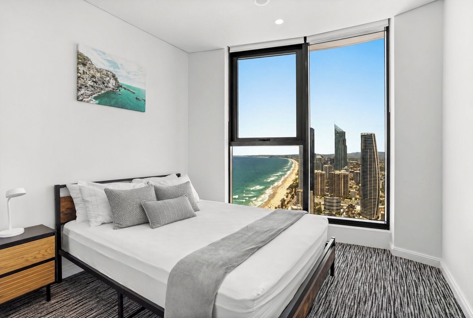 A modern bedroom with a large bed, a nightstand, a desk, and a window with a city and beach view.