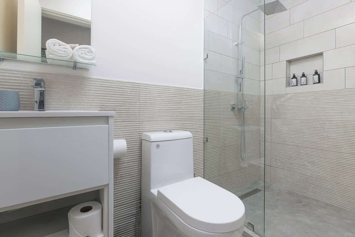 Uma casa de banho moderna com sanita, cabine de duche em vidro e uma prateleira com toalhas e produtos de higiene pessoal.