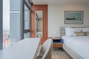 Habitación de hotel con cama, escritorio, silla, mesita de noche, lámpara, un cuadro y vistas de la ciudad.