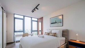 A modern hotel room with a large bed, a desk, a chair, a nightstand, a lamp, a window with a city view, and a framed picture on the wall. - Apartment Upper Level in Casco View (Panamá)