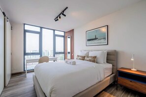 Ein modernes Hotelzimmer mit einem großen Bett, einem Schreibtisch, einem Stuhl, einem Nachttisch, einer Lampe, einem Fenster mit Blick auf die Stadt und einem gerahmten Bild an der Wand.