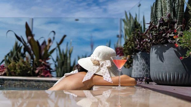 Una persona relajándose junto a una piscina con una bebida, rodeada de plantas y un cielo despejado.