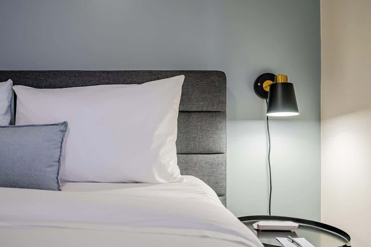 Un lit bien fait, avec des oreillers, une table de chevet ronde surmontée d’une lampe, et un luminaire fixé au mur.