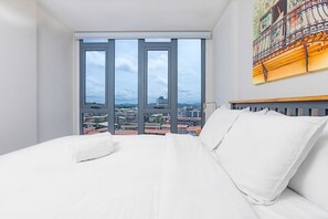 Une chambre d’hôtel avec un grand lit, du linge de lit blanc et une vue sur le paysage urbain grâce à de grandes fenêtres du sol au plafond.