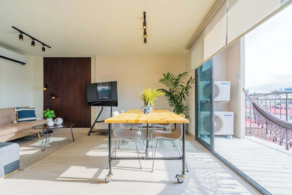 A modern living room with a sofa, a coffee table, a TV, a dining table with chairs, and a large window with a view.