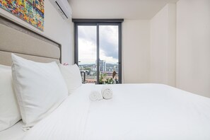 Habitación de hotel con una cama grande, ropa de cama blanca, vistas a la ciudad a través de una puerta corredera y aire acondicionado instalado en el techo.