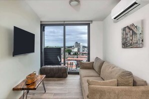 Ein modernes Wohnzimmer mit einer Couch, einem Flachbildfernseher, einem Holztisch und einer Glastür, die auf einen Balkon führt.