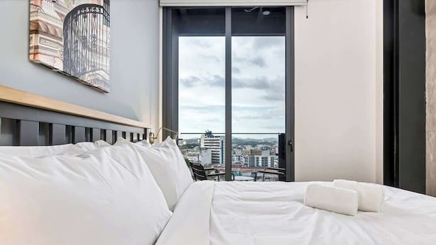 Hotellihuone, jossa on suuri sänky, liukuoven kautta avautuva näkymä kaupunkiin ja seinällä oleva kehystetty taideteos.