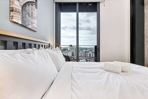 Ein Hotelzimmer mit einem großen Bett, Blick auf die Stadt durch eine Schiebetür und einem gerahmten Kunstwerk an der Wand.