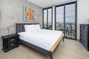 Ein Schlafzimmer mit einem großen Bett, einem Nachttisch, einer Kommode und Blick auf die Stadt durch große Fenster.