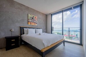 A modern bedroom with a large bed, a nightstand, and a view of a cityscape through a large window. - Penthouse with Amador bay views and dual balconies (Panama City)