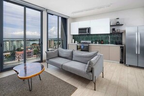 Eine moderne Wohnung mit grauem Sofa, einem kleinen Holztisch, einer Küche mit weißen Schränken und einem großen Fenster mit Blick auf die Stadt.