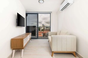 A modern living room with a flat-screen TV, a sofa, a wooden console table, and a sliding glass door leading to a balcony with a city view. - Jr. Apartment Upper Level in Casco View (Panama City)
