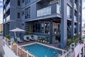 Ein modernes, mehrgeschossiges Gebäude mit Pool auf dem Dach, Außenbereich zum Sitzen und einer überdachten Fläche mit Pflanzen.